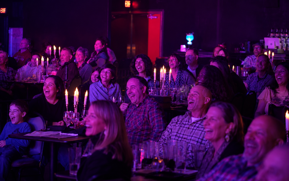Audience laughing and clapping in a dimly lit venue with candlelit tables.