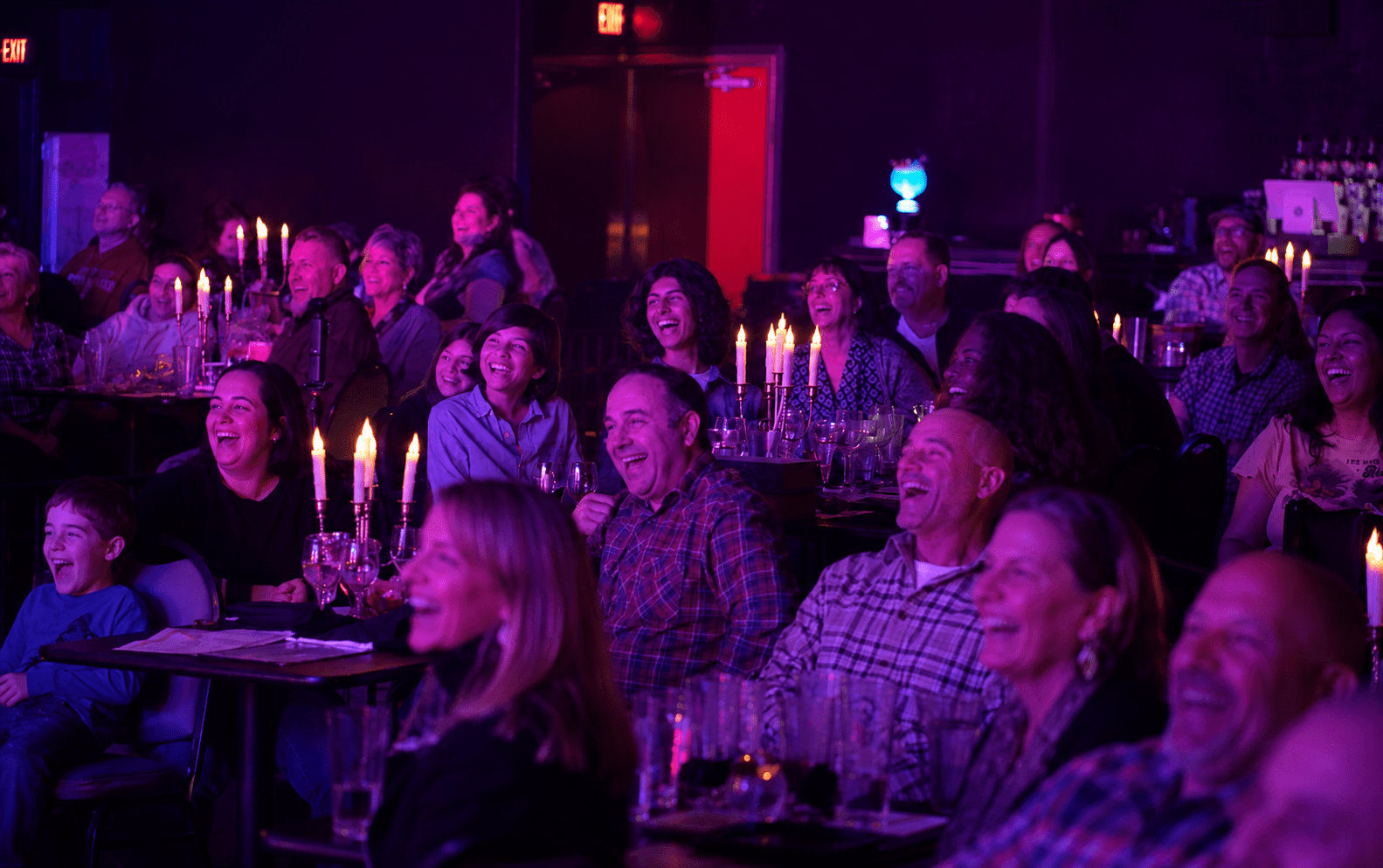 Audience laughing and clapping in a dimly lit venue with candlelit tables.