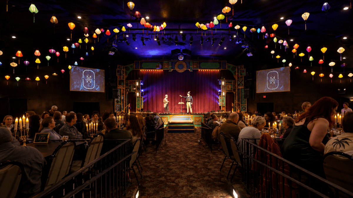 Two performers on stage, colorful lanterns overhead, audience seated in rows.