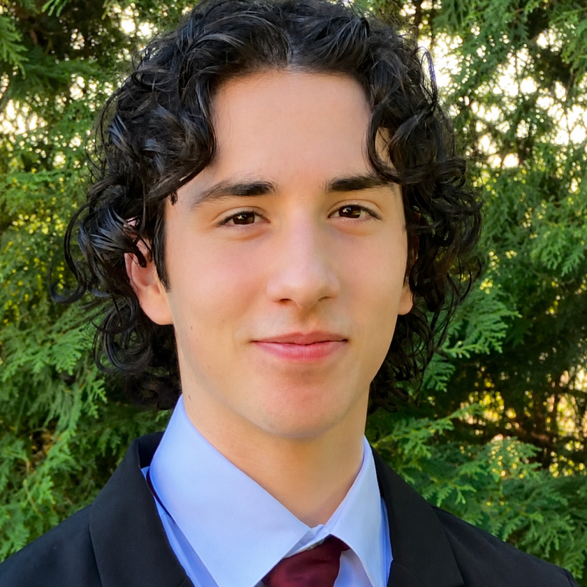 Smiling person in suit and tie with curly hair, standing outdoors.