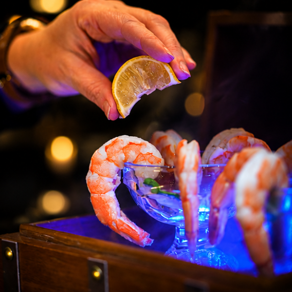 Hand squeezing lemon over shrimp cocktail in blue-lit dish.