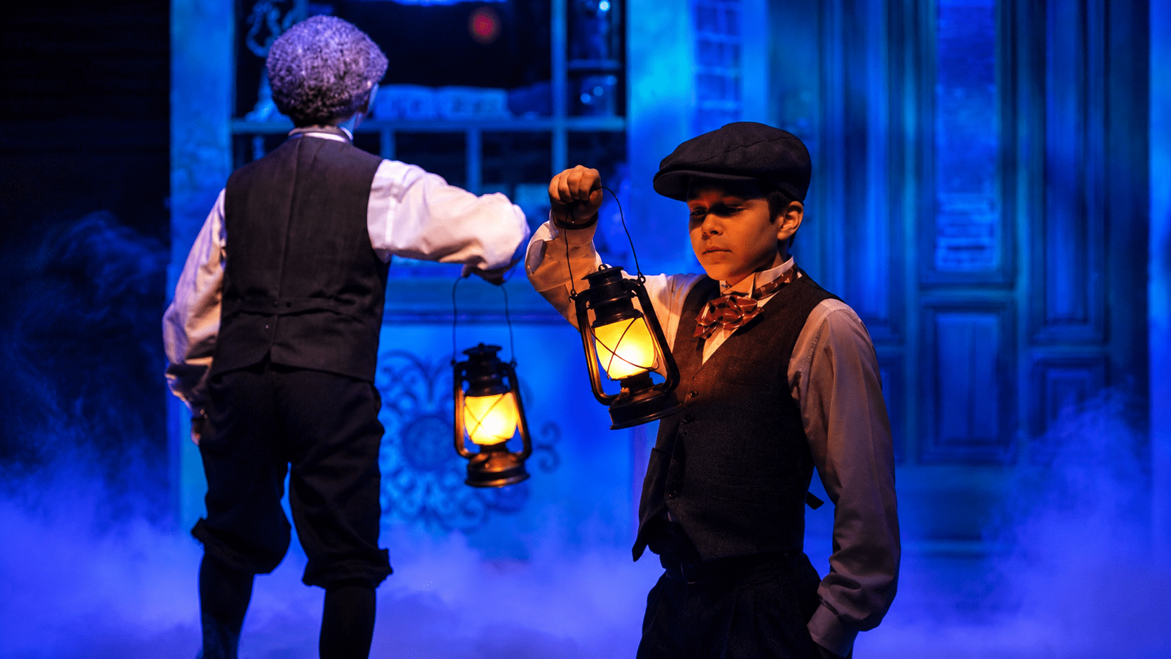 Two children in vintage attire hold lanterns on a foggy stage.