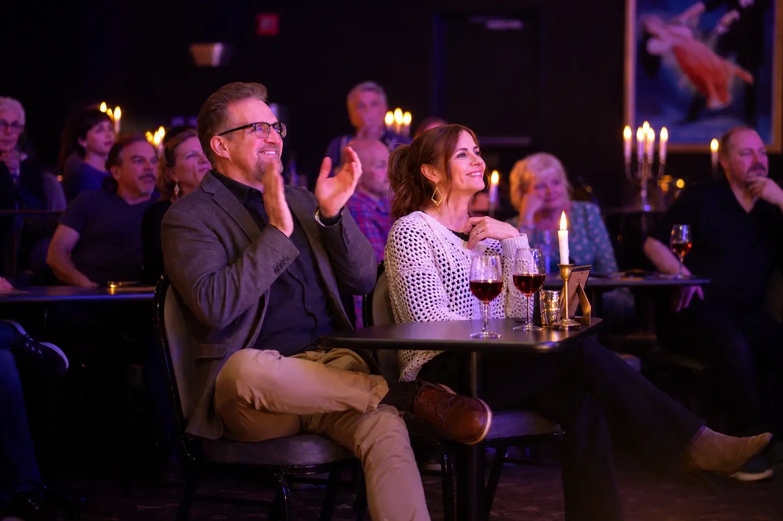 Audience seated at tables, clapping with drinks, in a dimly lit venue.