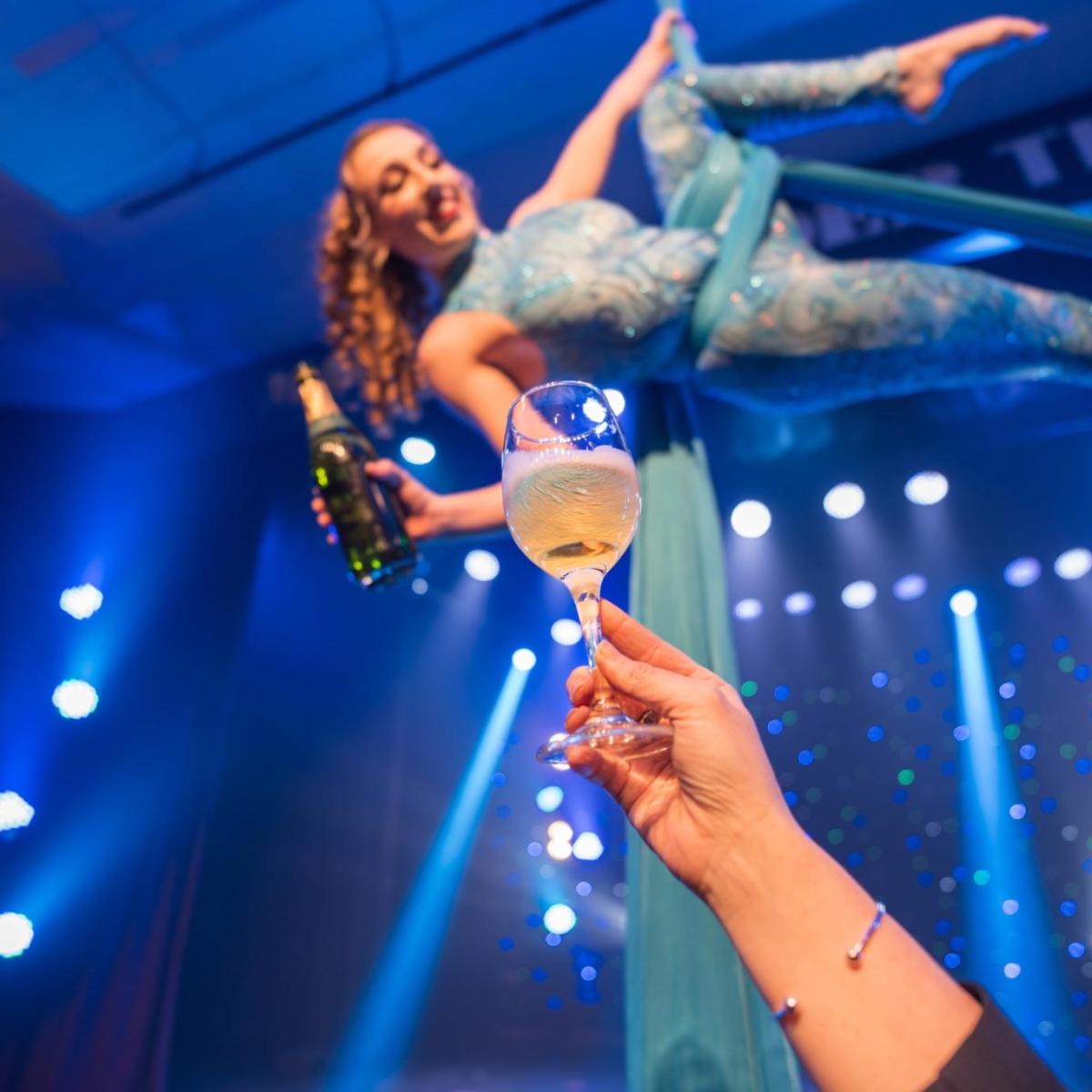 Aerial performer pouring champagne into a glass on stage with colorful lights.