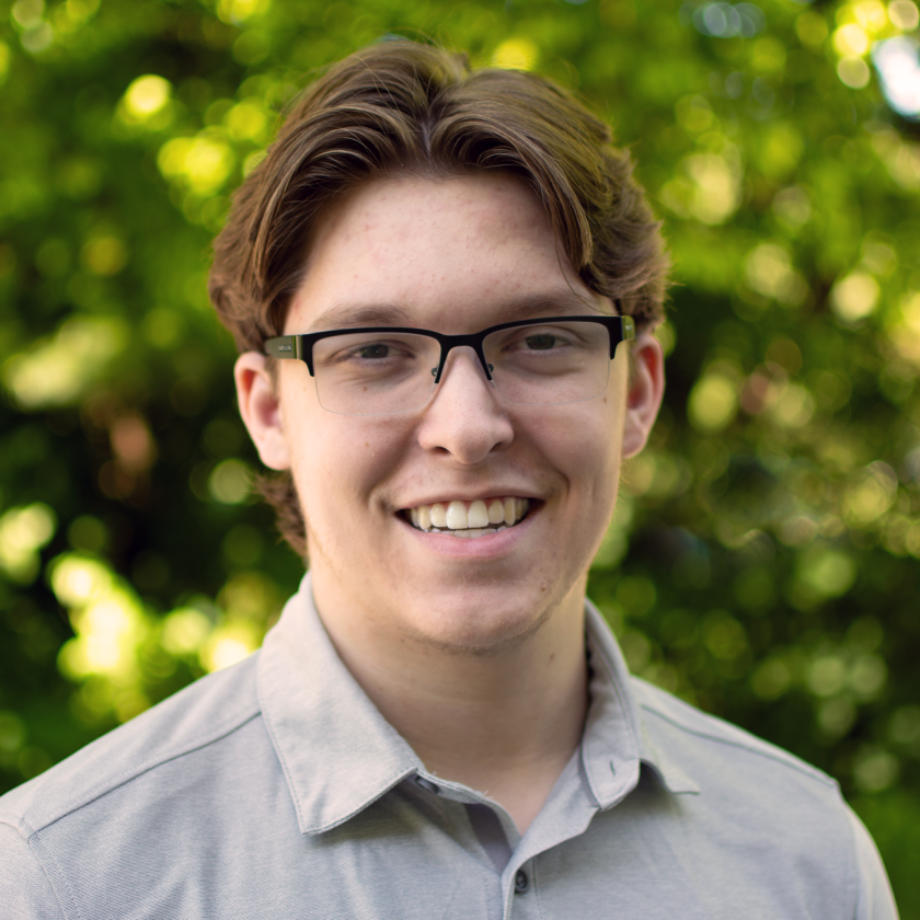 Person with glasses smiling in front of leafy background.