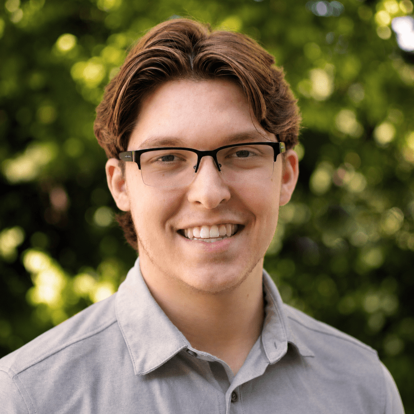 Person with glasses smiling in front of leafy background.