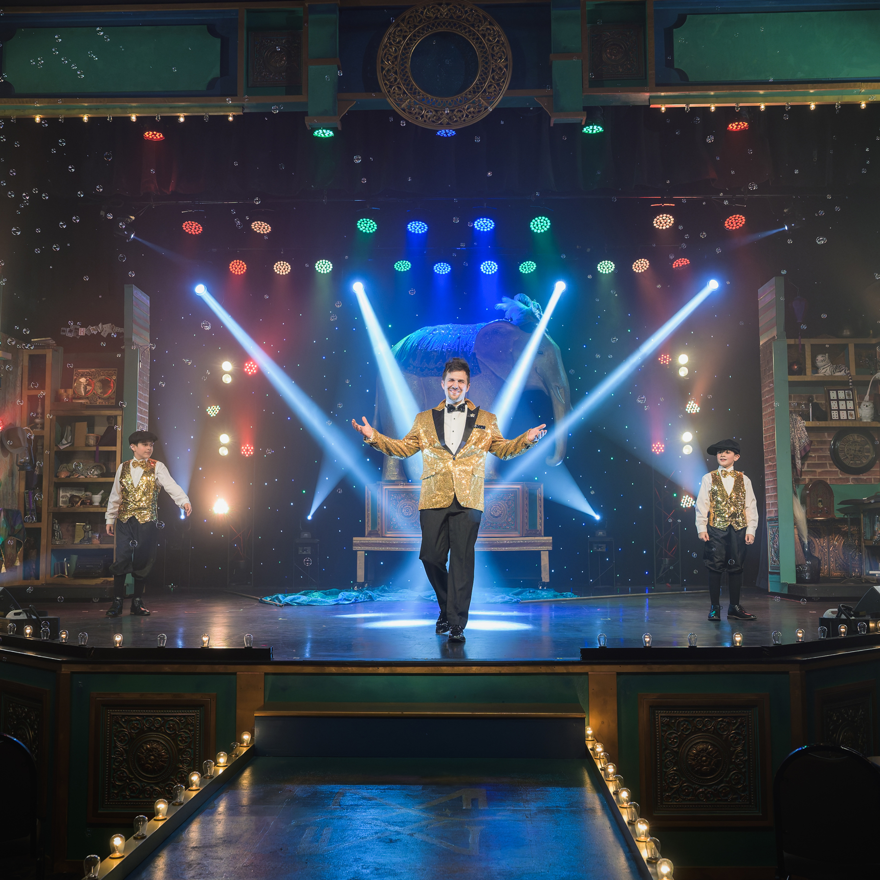 Performers in gold jackets with stage lights and bubbles, on a colorful theater stage.