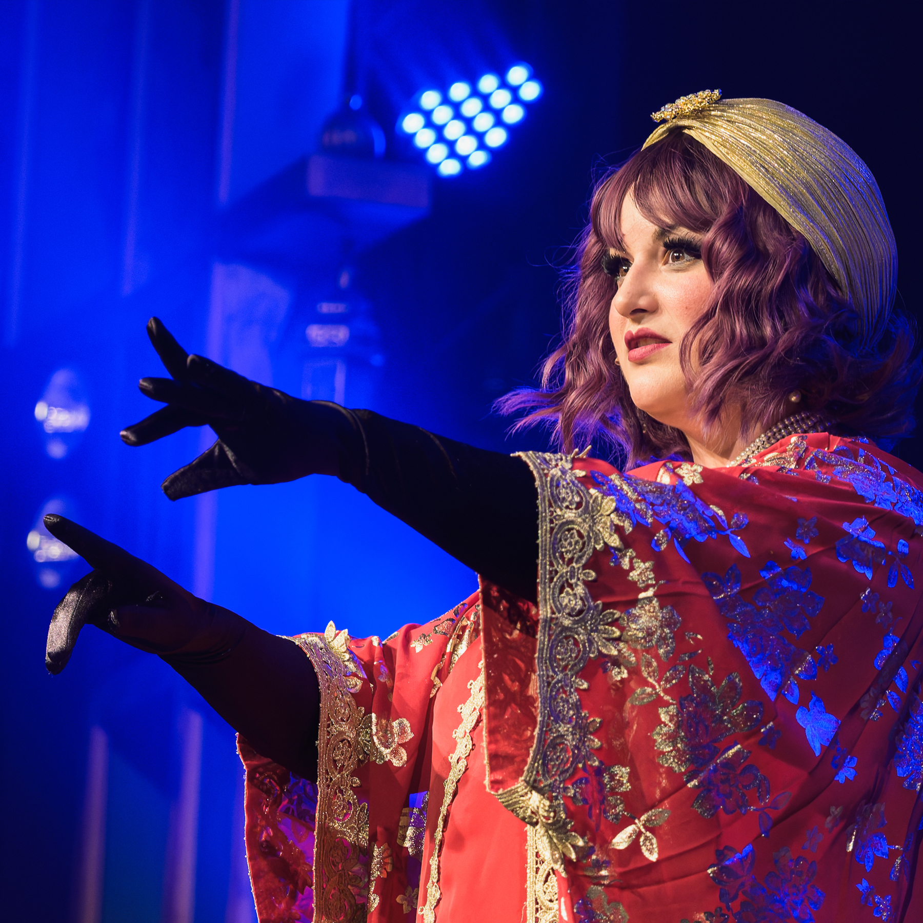 Performer in ornate costume with raised hand on stage, blue lighting.