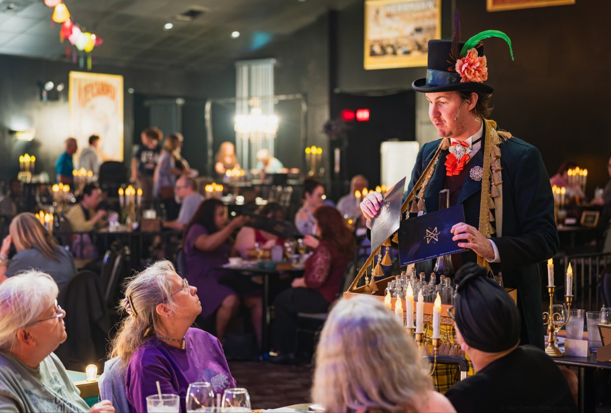 A costumed man with a top hat engages with seated patrons in a dimly lit restaurant.