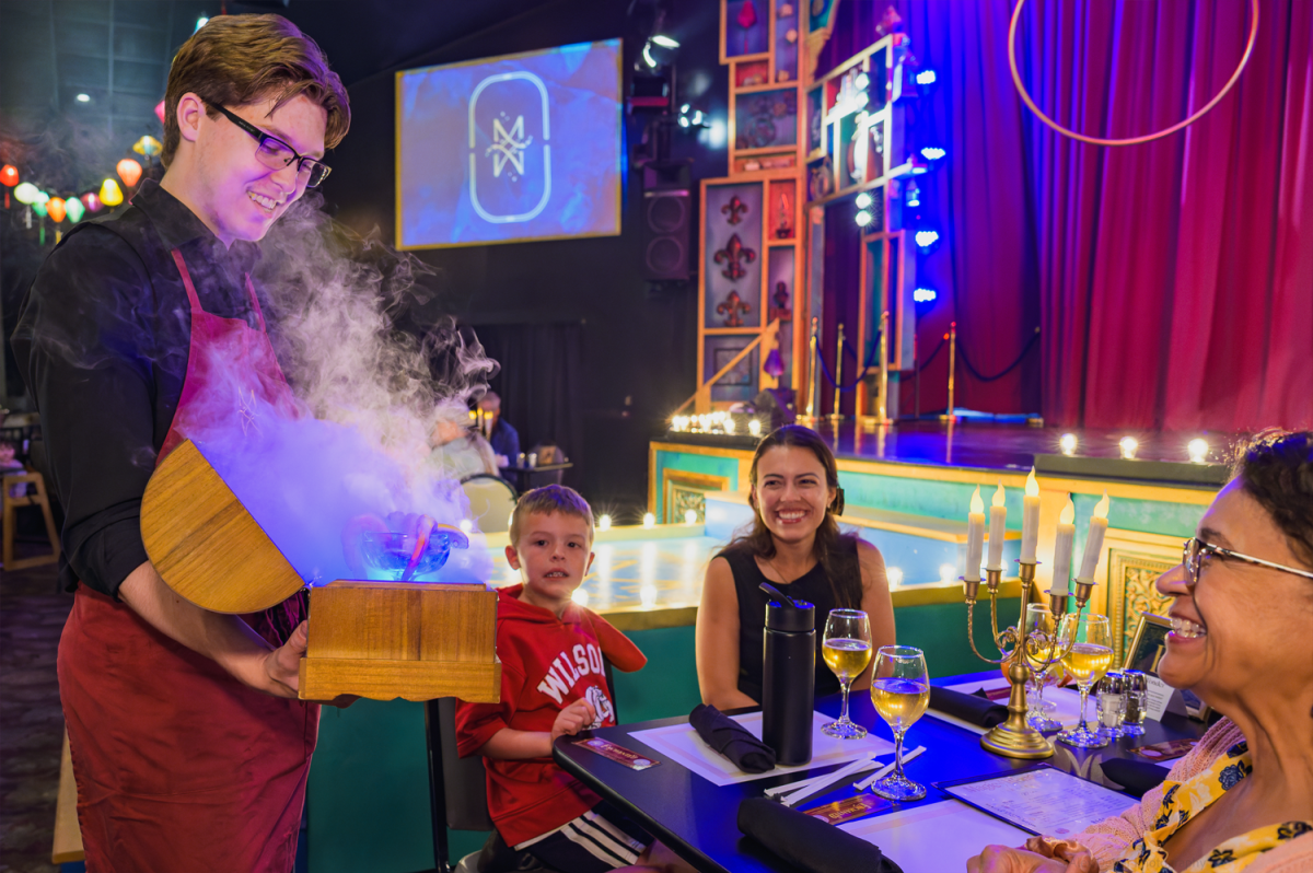 Server presents steaming dish to smiling diners in a colorful, theatrical restaurant setting.