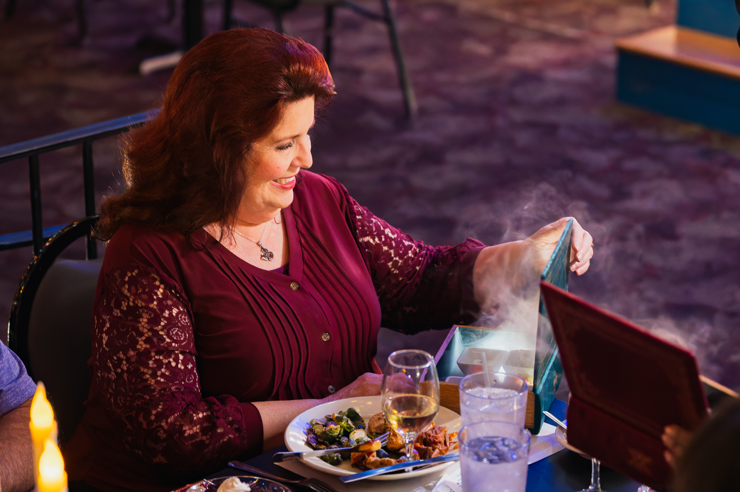 Woman opening steaming gift box at restaurant with meal on table.