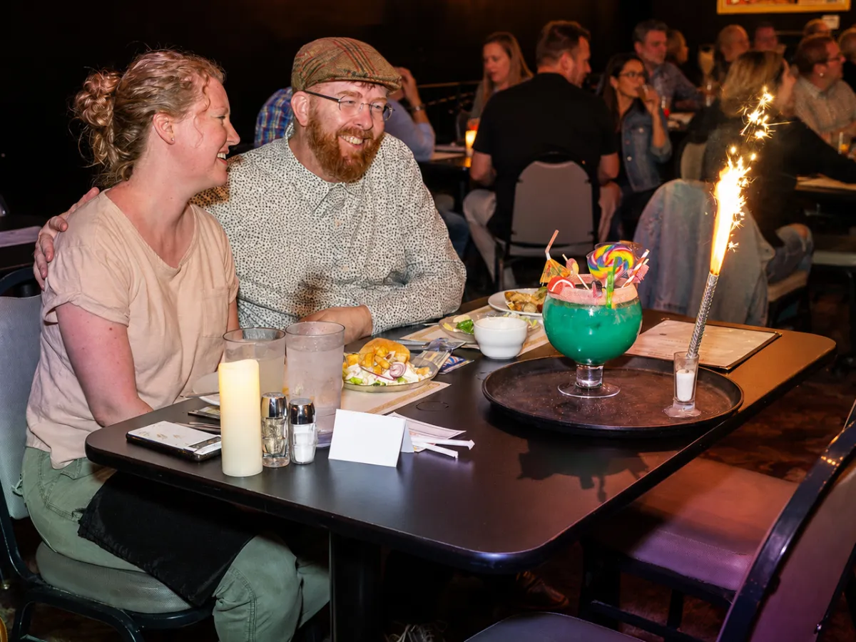 Smiling couple at a restaurant table with a large, vibrant cocktail and sparkler.