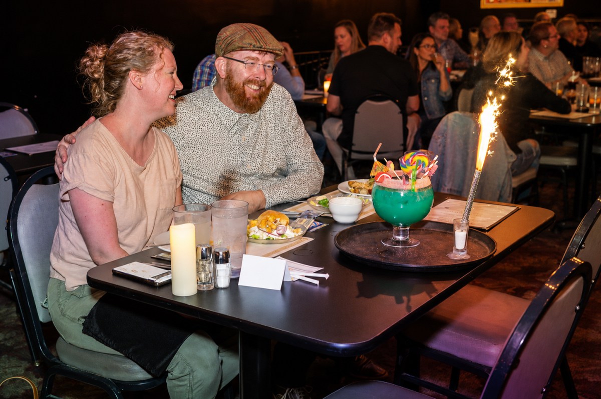Smiling couple at a restaurant table with a large, vibrant cocktail and sparkler.